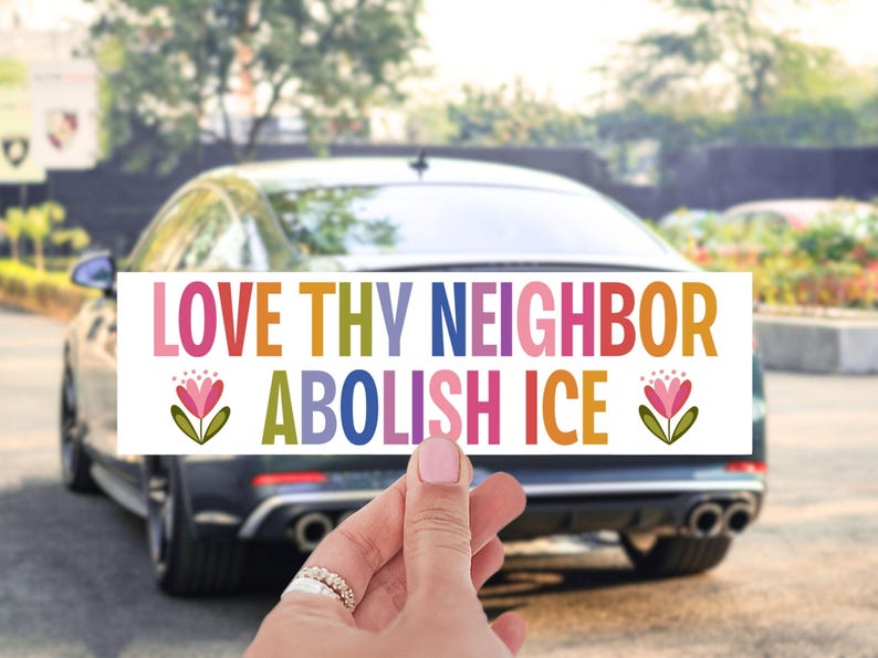 May include: A rectangular sticker with the text "LOVE THY NEIGHBOR ABOLISH ICE" in colorful block letters. The sticker is held in front of a car. Floral illustrations are on the sticker.