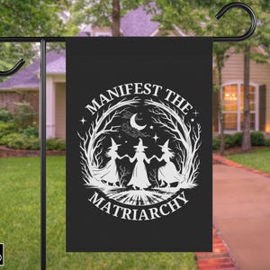 May include: Black garden flag with a white graphic of three witches holding hands, surrounded by a tree and crescent moon. The text reads "Manifest the Matriarchy". The flag is hanging on a black metal stand.