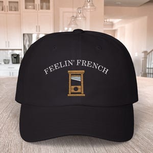 May include: A black baseball cap with white embroidery that reads "Feelin' French" and a small embroidered image of a guillotine.
