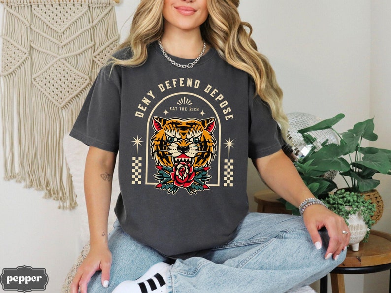 May include: A dark gray t-shirt with a graphic of a tiger's face in a vintage tattoo style. The text "Deny Defend Depose Eat The Rich" is printed around the tiger's face.