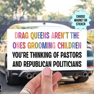 Op de afbeelding: Een witte rechthoekige magneet of sticker met de tekst "DRAG QUEENS AREN'T THE ONES GROOMING CHILDREN YOU'RE THINKING OF PASTORS AND REPUBLICAN POLITICIANS" in kleurrijke letters. De achtergrond toont een auto en een vage buitensc&egrave;ne.