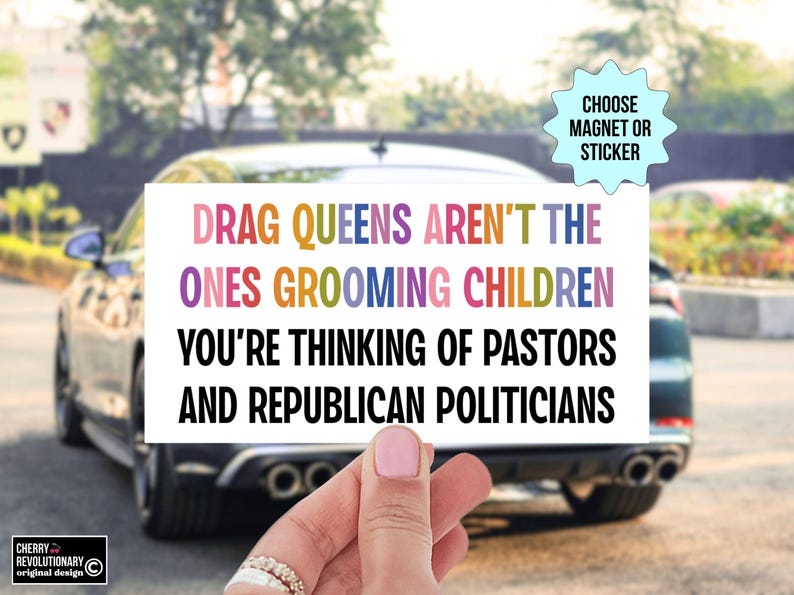 Op de afbeelding: Een witte rechthoekige magneet of sticker met de tekst "DRAG QUEENS AREN'T THE ONES GROOMING CHILDREN YOU'RE THINKING OF PASTORS AND REPUBLICAN POLITICIANS" in kleurrijke letters. De achtergrond toont een auto en een vage buitensc&egrave;ne.