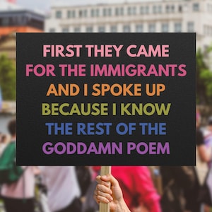 May include: A black sign with the text "FIRST THEY CAME FOR THE IMMIGRANTS AND I SPOKE UP BECAUSE I KNOW THE REST OF THE GODDAMN POEM" in rainbow colors. The sign is held up in a crowd.