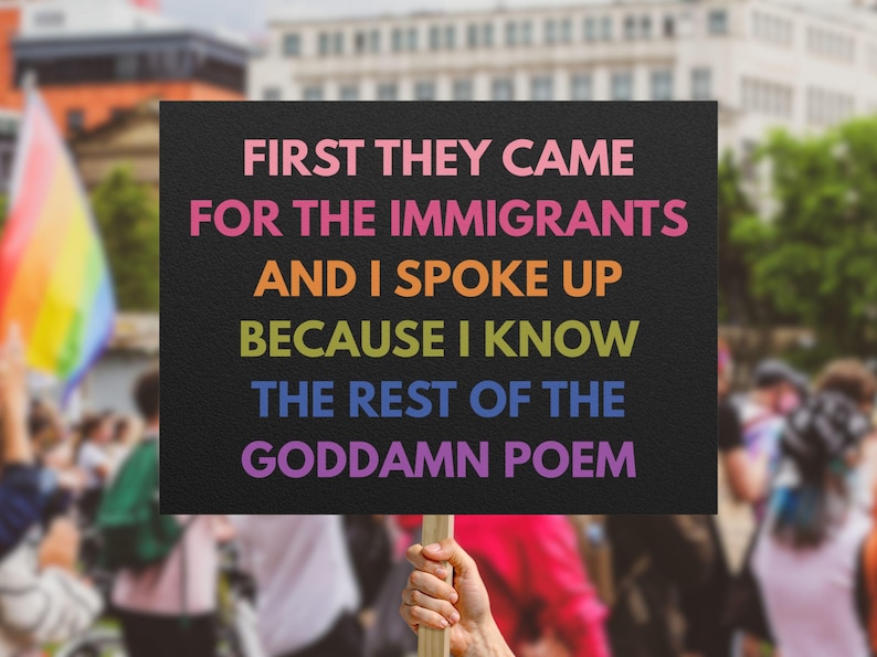 May include: A black sign with the text "FIRST THEY CAME FOR THE IMMIGRANTS AND I SPOKE UP BECAUSE I KNOW THE REST OF THE GODDAMN POEM" in rainbow colors. The sign is held up in a crowd.