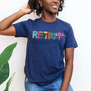 Puede incluir: Una camiseta azul marino con la palabra "RESIST" escrita en letras de colores. Cada letra est&aacute; decorada con un s&iacute;mbolo diferente, incluyendo un pu&ntilde;o, una bandera arco&iacute;ris, un &uacute;tero y un &aacute;tomo.