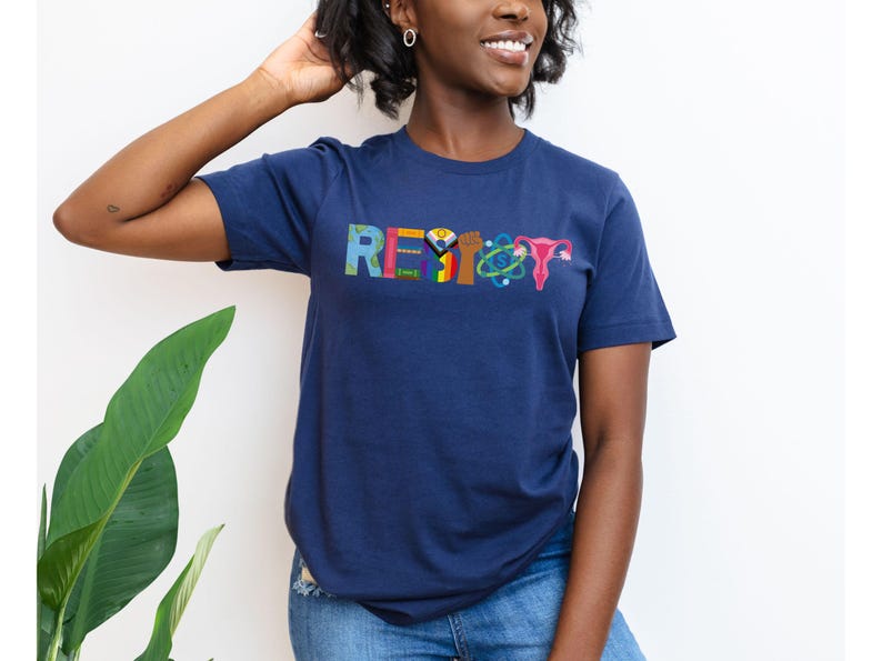 Puede incluir: Una camiseta azul marino con la palabra "RESIST" escrita en letras de colores. Cada letra est&aacute; decorada con un s&iacute;mbolo diferente, incluyendo un pu&ntilde;o, una bandera arco&iacute;ris, un &uacute;tero y un &aacute;tomo.