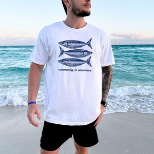 May include: White t-shirt with a graphic of three blue fish and the text "community is resistance". The person wearing the shirt is standing on a beach.