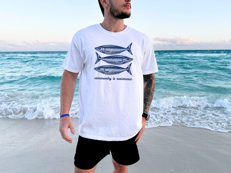 May include: White t-shirt with a graphic of three blue fish and the text "community is resistance". The person wearing the shirt is standing on a beach.