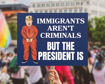 May include: A protest sign held up in a crowd. The sign is dark blue with white text that reads "IMMIGRANTS AREN'T CRIMINALS BUT THE PRESIDENT IS." An illustration of a person in an orange jumpsuit is on the sign.