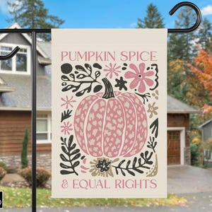 Può includere: Una bandiera da giardino color crema con un disegno di zucca rosa e le parole "PUMPKIN SPICE & EQUAL RIGHTS". La bandiera è decorata con motivi floreali e di foglie in nero, rosa e marrone. La bandiera è appesa a un supporto di metallo nero.