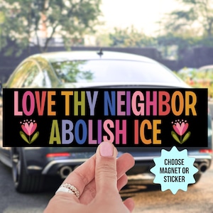 May include: A black rectangular bumper sticker with the words "LOVE THY NEIGHBOR ABOLISH ICE" in colorful letters. The sticker features floral accents and is held in front of a car.