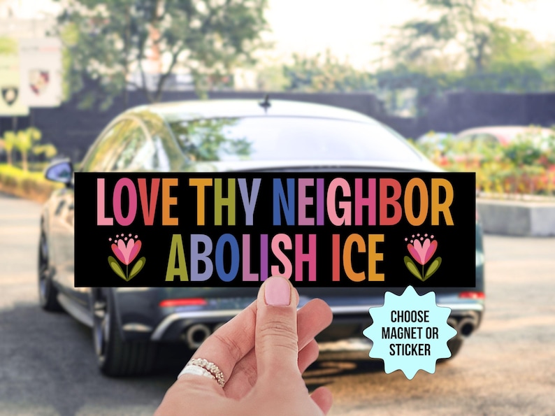 May include: A black rectangular bumper sticker with the words "LOVE THY NEIGHBOR ABOLISH ICE" in colorful letters. The sticker features floral accents and is held in front of a car.