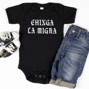 May include: A black baby onesie with the text "CHINGA LA MIGRA" in white gothic lettering, paired with blue denim jeans and a pair of black and white baby sneakers. The items are arranged on a white surface.