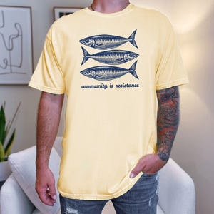May include: A yellow t-shirt with a graphic of three fish and the text "community is resistance".