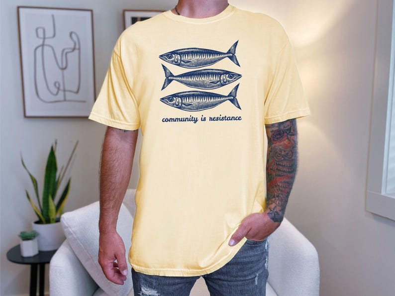 May include: A yellow t-shirt with a graphic of three fish and the text "community is resistance".