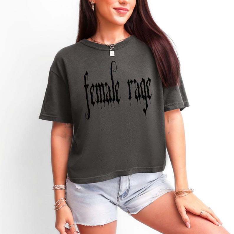 May include: Dark grey t-shirt with the words "female rage" in a gothic font. The shirt has short sleeves and a relaxed fit. The text is in black.