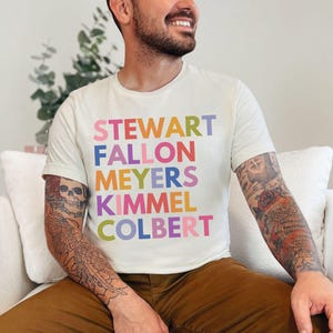 May include: Cream-colored t-shirt with the names STEWART, FALLON, MEYERS, KIMMEL, and COLBERT in colorful block letters. The person wearing the shirt is seated and has tattoos on their arms.