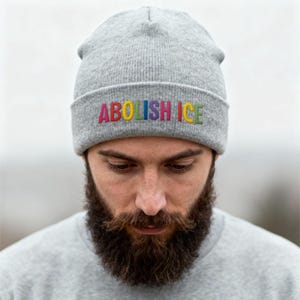 May include: A gray beanie with the embroidered text "ABOLISH ICE" in rainbow colors. The beanie is made of a soft, knitted material. The person wearing the beanie has a full beard and is wearing a gray shirt.