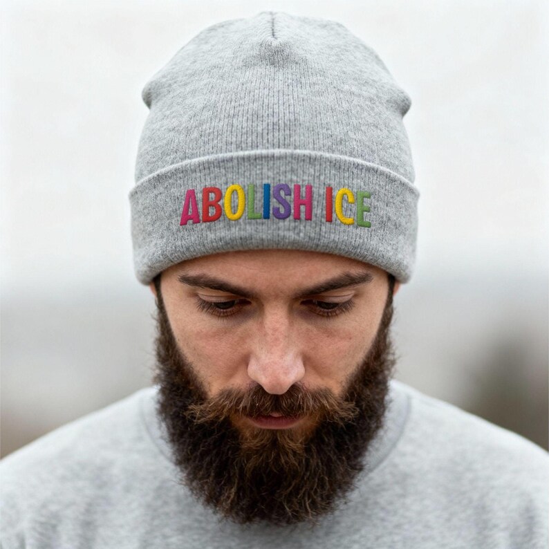 May include: A gray beanie with the embroidered text "ABOLISH ICE" in rainbow colors. The beanie is made of a soft, knitted material. The person wearing the beanie has a full beard and is wearing a gray shirt.