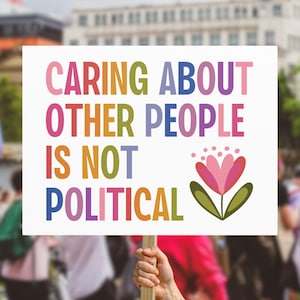 May include: A white sign held up in a crowd with the words "CARING ABOUT OTHER PEOPLE IS NOT POLITICAL" in colorful letters. A pink and purple flower graphic is on the right side of the sign.