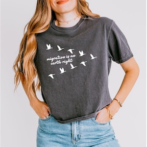 May include: Dark gray t-shirt with the text "migration is an earth right" and a flock of white birds in flight. The shirt is a short-sleeved, crew neck style.