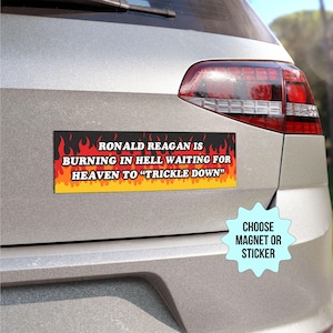 May include: A black and orange bumper sticker with the text "RONALD REAGAN IS BURNING IN HELL WAITING FOR HEAVEN TO "TRICKLE DOWN"" on a silver car. The sticker has flames on the sides. A blue starburst sticker with the text "CHOOSE MAGNET OR STICKER" is on the car.