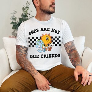 May include: A white t-shirt with a graphic design featuring a sun and a planet with the text "COPS ARE NOT OUR FRIENDS" in black letters.