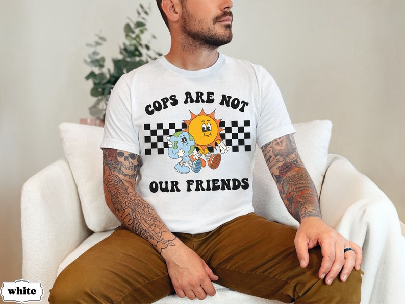 May include: A white t-shirt with a graphic design featuring a sun and a planet with the text "COPS ARE NOT OUR FRIENDS" in black letters.
