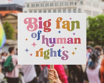 May include: A white sign with the text "Big fan of human rights" in colorful, retro-style lettering. The sign is held up in a crowd, with people and buildings in the background. Decorative stars surround the text.