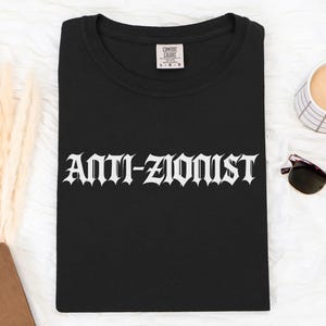 Puede incluir: Una camiseta negra con el texto ANTI-ZIONIST impreso en una fuente blanca y audaz de estilo gótico en el pecho. La prenda tiene un cuello redondo y una etiqueta de la marca Comfort Colors visible en la zona interior del cuello.