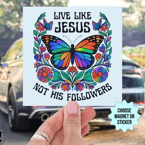 May include: A square sticker or magnet featuring a colorful butterfly with rainbow wings, surrounded by flowers and the text "LIVE LIKE JESUS NOT HIS FOLLOWERS." The design is held in front of a car.