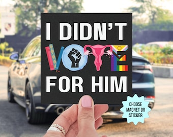 May include: Black and white square sign with the text "I didn't vote for him" in white. The word "vote" is spelled out with various symbols, including a book, a fist, a uterus, and a rainbow flag.