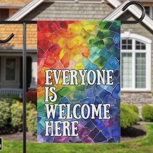 May include: A garden flag with the text "EVERYONE IS WELCOME HERE" in white block letters. The background is a mosaic of colorful, stained-glass-like shapes in red, orange, yellow, green, blue, and purple. The flag is hanging on a black metal pole.