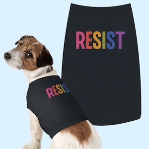 May include: A small dog wearing a black dog shirt with the word "RESIST" in rainbow colors. A matching shirt is displayed next to the dog. The dog has brown and white fur and is sitting on a light blue background.
