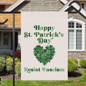 Pode incluir: Uma bandeira de jardim do Dia de São Patrício com o texto "Happy St. Patrick's Day" em verde, um coração feito de trevos verdes e as palavras "Resist Fascism". A bandeira é exibida em um suporte de metal preto.