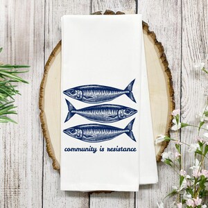 May include: White kitchen towel with three blue fish illustrations and the phrase "community is resistance." The fish are stylized with detailed scales and fins. The towel is folded and displayed on a wooden surface.