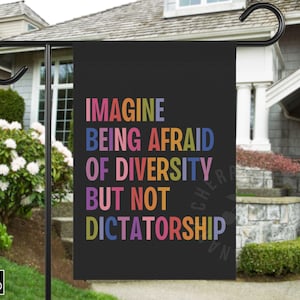 Op de afbeelding: Zwarte tuinvaandel met de tekst "IMAGINE BEING AFRAID OF DIVERSITY BUT NOT DICTATORSHIP" in kleurrijke letters. De vlag wordt buiten op een zwarte metalen standaard voor een huis getoond.
