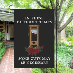 May include: Black garden flag with the text "IN THESE DIFFICULT TIMES" and "SOME CUTS MAY BE NECESSARY". The flag features an illustration of a guillotine with red roses at the bottom. The flag is hanging in a garden.