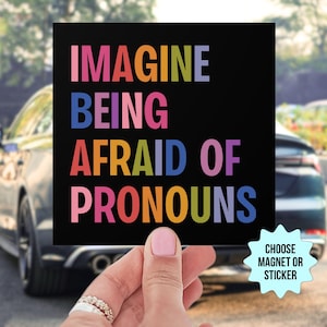 Può includere: Un magnete o un adesivo quadrato nero con il testo "Imagine being afraid of pronouns" in colori arcobaleno. Il testo è in maiuscolo. Il magnete o l'adesivo è tenuto da una mano.