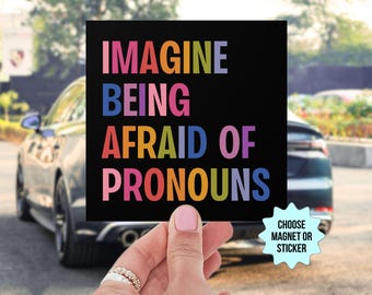 May include: A black square magnet or sticker with the text "Imagine being afraid of pronouns" in rainbow colors. The text is in all caps. The magnet or sticker is being held by a hand.