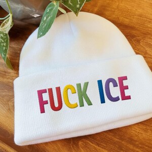 May include: A white beanie with the embroidered text "FUCK ICE" in rainbow colors. The hat is made of a soft, knitted material and is suitable for cold weather. The beanie is a casual accessory.