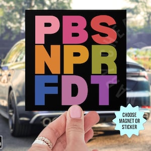 May include: A black square magnet or sticker with the text "PBS NPR FDT" in colorful block letters. The words are stacked vertically. The background shows a car and a hand holding the magnet.