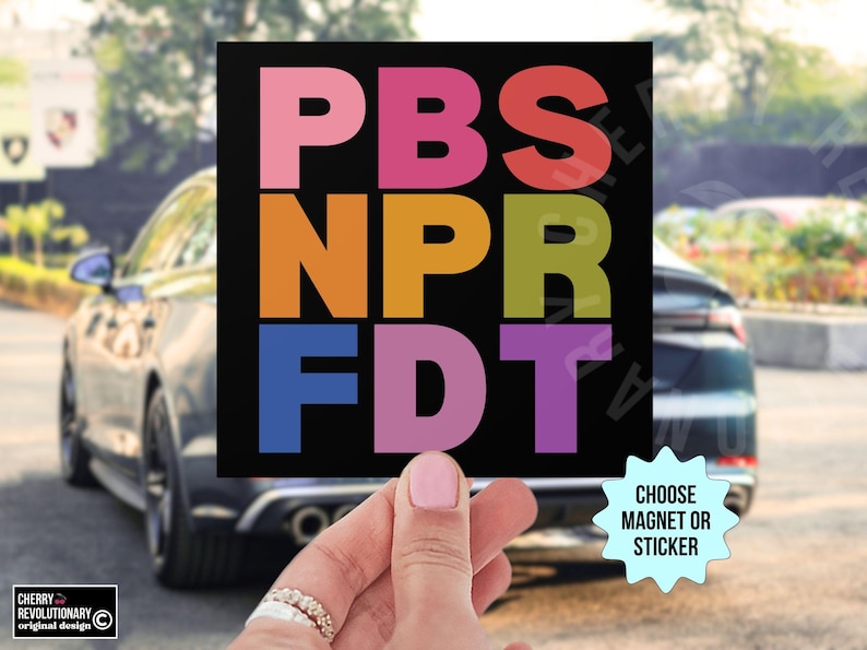 May include: A black square magnet or sticker with the text "PBS NPR FDT" in colorful block letters. The words are stacked vertically. The background shows a car and a hand holding the magnet.