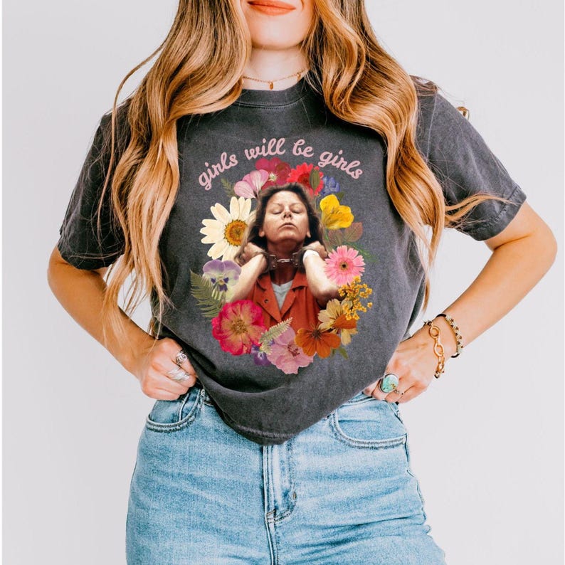 May include: Dark gray t-shirt featuring a floral design surrounding an image of a person. The text "girls will be girls" is arched above the image. The shirt is worn by a person wearing blue jeans.