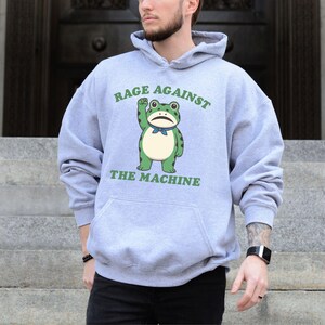 May include: A light gray hoodie with a front graphic of a green frog with a raised fist and the text "RAGE AGAINST THE MACHINE" in green. The hoodie has a kangaroo pocket and a drawstring hood.