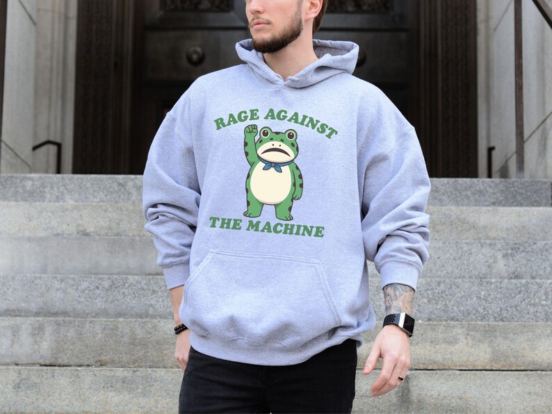 May include: A light gray hoodie with a front graphic of a green frog with a raised fist and the text "RAGE AGAINST THE MACHINE" in green. The hoodie has a kangaroo pocket and a drawstring hood.