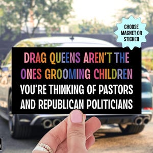 Op de afbeelding: Een zwarte rechthoekige magneet of sticker met de tekst "DRAG QUEENS AREN'T THE ONES GROOMING CHILDREN YOU'RE THINKING OF PASTORS AND REPUBLICAN POLITICIANS" in kleurrijke letters. De magneet wordt voor een auto gehouden.