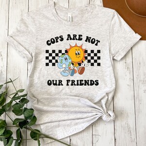 May include: A gray t-shirt with a graphic of a sun and earth holding hands. The text "COPS ARE NOT OUR FRIENDS" is printed in black letters on the shirt.