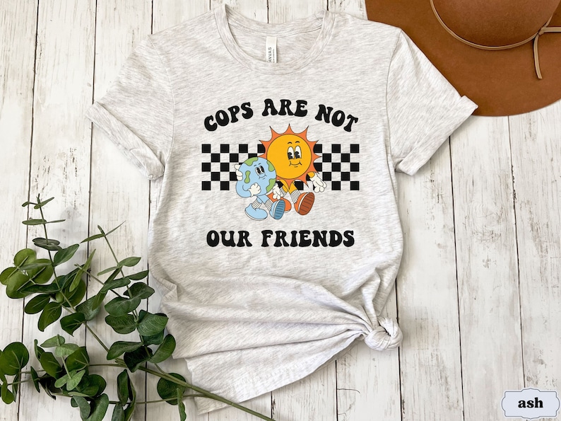 May include: A gray t-shirt with a graphic of a sun and earth holding hands. The text "COPS ARE NOT OUR FRIENDS" is printed in black letters on the shirt.