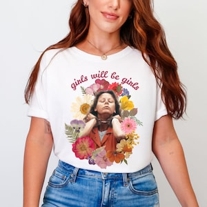 May include: White t-shirt with the text "girls will be girls" in a cursive font. The shirt features a graphic of a person surrounded by colorful flowers. The person is wearing an orange jacket.